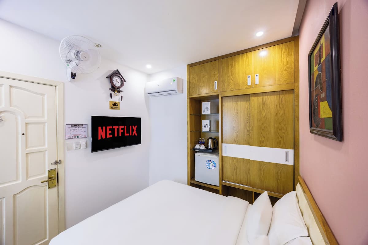 A hotel room with a bed, a wardrobe, a small refrigerator, a clock, an air conditioner, a wall-mounted fan, and a Netflix sign.