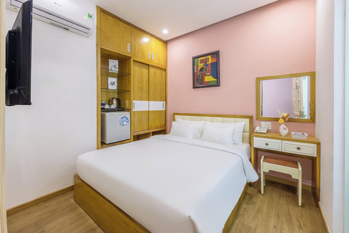 A hotel room with a bed, a desk, a wardrobe, a small fridge, a mirror, and a painting on the wall.