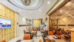 A hotel dining area with orange chairs, tables, a TV screen, and a large mirror.