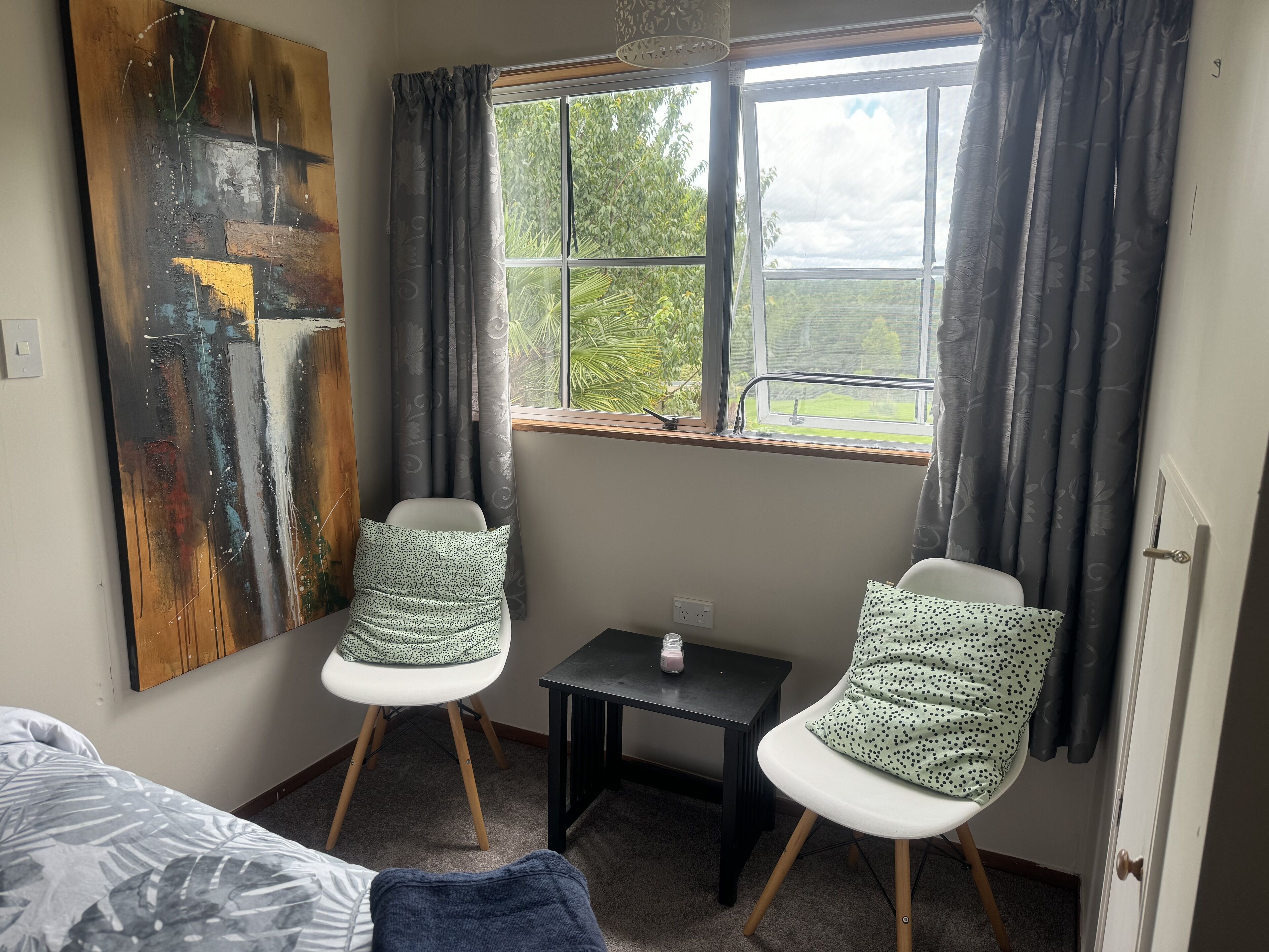 A room with two white chairs, a small table, a bed, and a large abstract painting on the wall.
