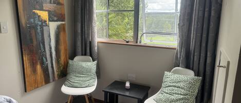 A room with two white chairs, a small table, a bed, and a large abstract painting on the wall.