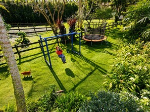 Een kind speelt op een schommel in een verzorgde tuin, met een trampoline en een hond in de buurt.