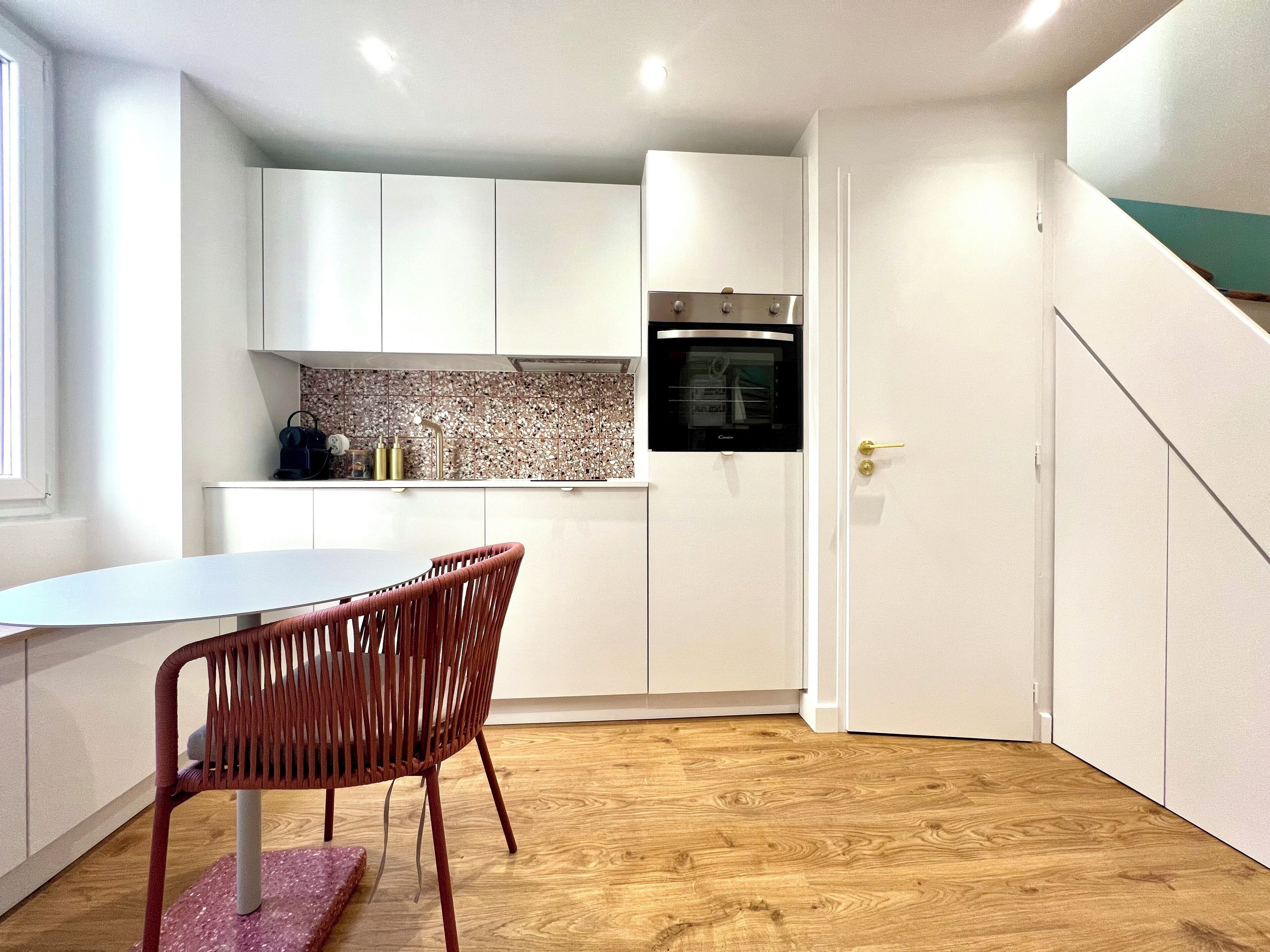 Een compacte keuken met witte kastjes, een inbouwoven en een eethoek met een tafel en stoelen.