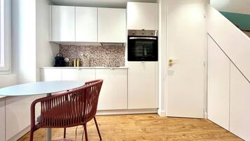 Een compacte keuken met witte kastjes, een inbouwoven en een eethoek met een tafel en stoelen.
