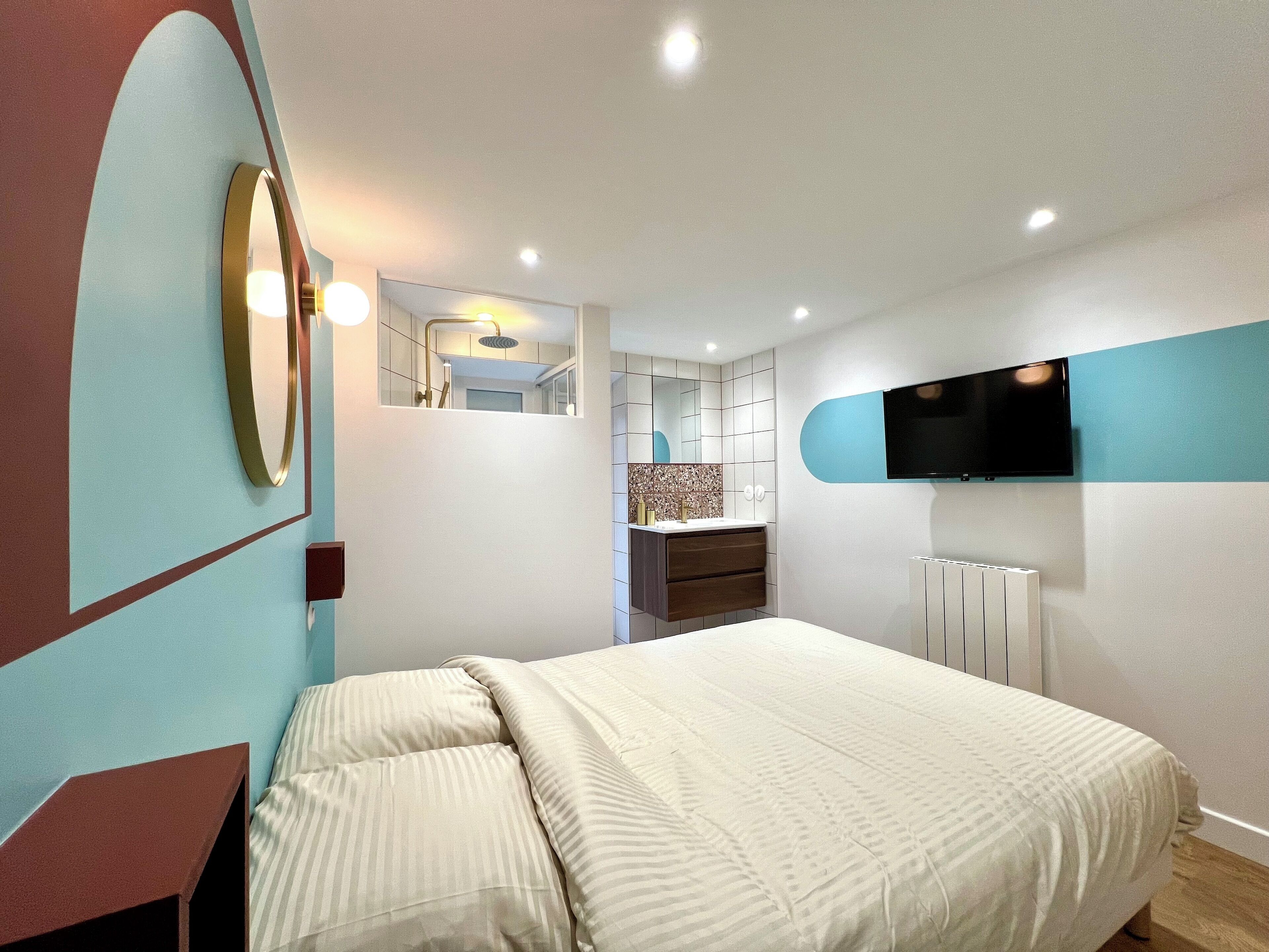 Un dormitorio moderno con una cama grande, un televisor en la pared y un baño a la vista a través de una puerta abierta.