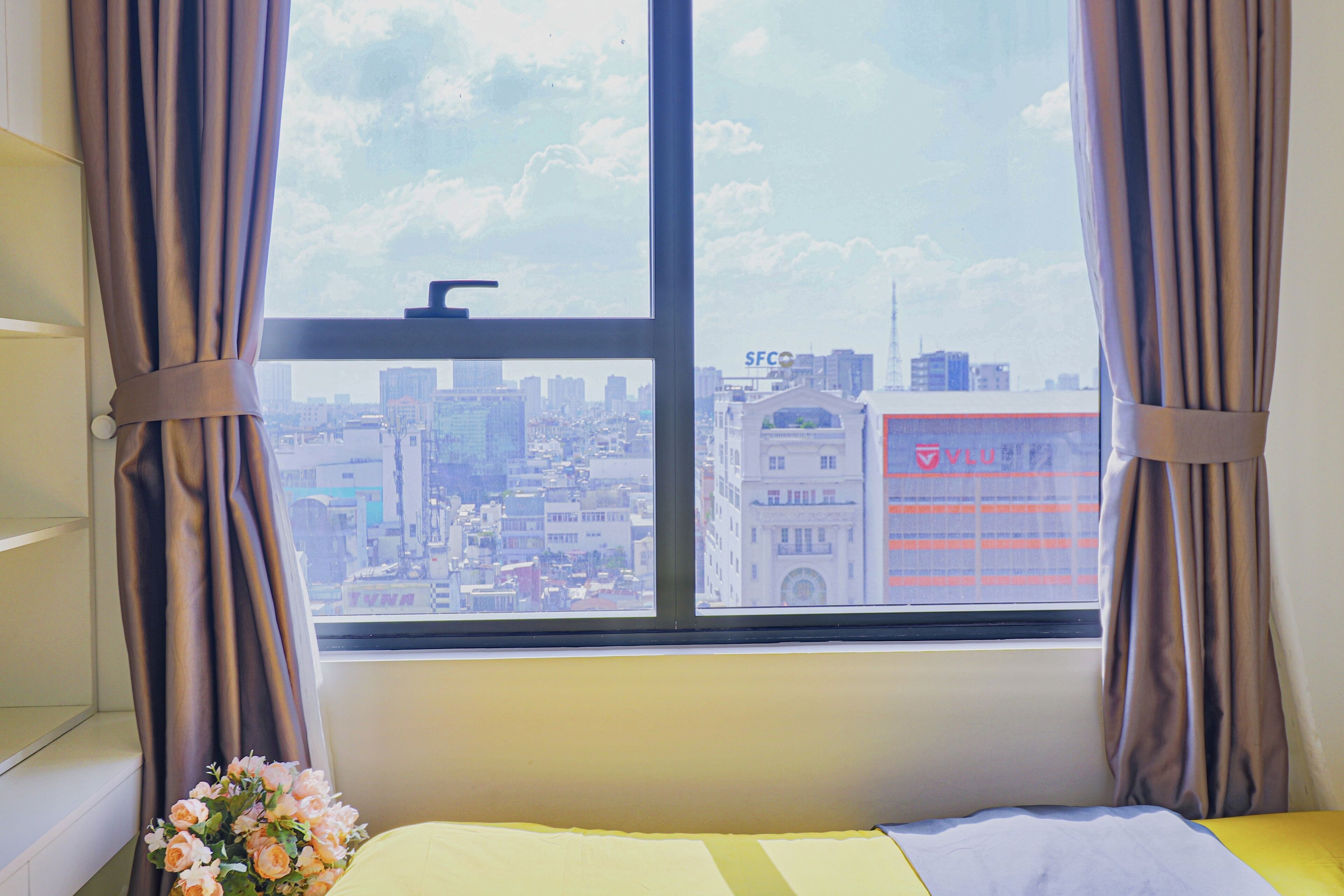 A room with a large window, curtains, a bed, and a vase with flowers.