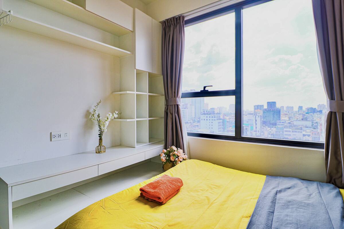 Ein Hotelzimmer mit Bett, Fenster mit Stadtblick, einem weißen Regal und einer Blumenvase.
