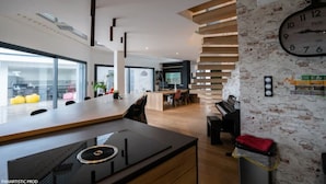A modern kitchen with a built-in stove, a dining area with a long table, and a brick wall with a clock.