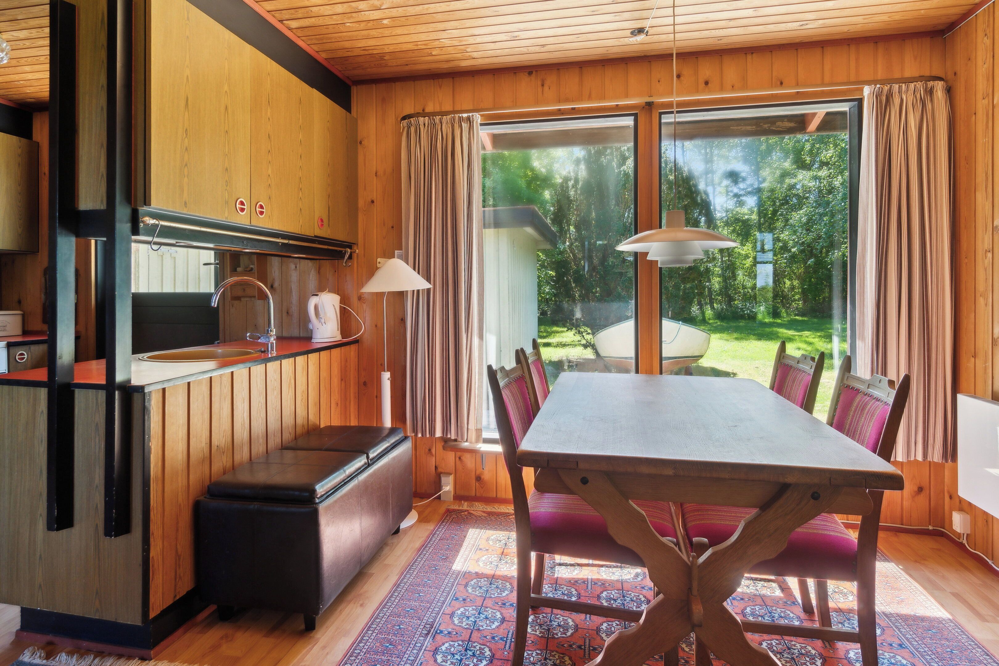 A wooden interior with a dining table, chairs, a kitchen area, and a large window.