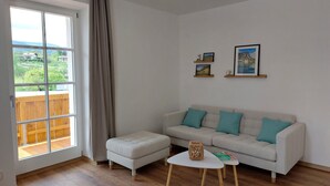 Un salon doté d’une grande fenêtre, d’un canapé gris agrémenté de coussins turquoise, d’une petite table blanche et d’un parquet en bois.