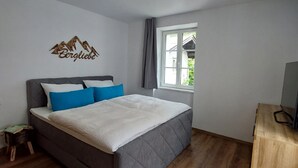 Une chambre à coucher avec un lit, un petit tabouret en bois, une fenêtre avec des rideaux et un mur décoré d’un motif de montagne.