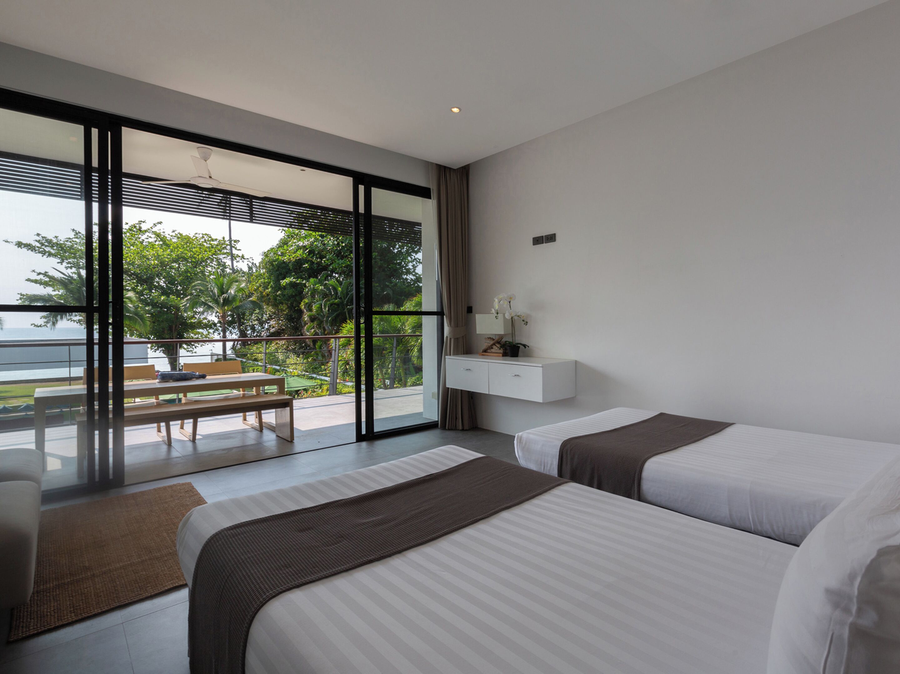 A modern hotel room with two beds, a small desk, and a balcony with a view of the sea.