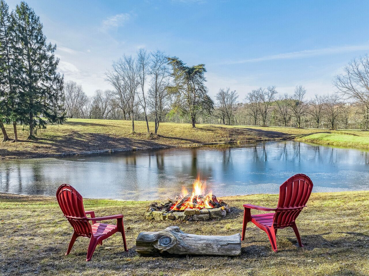 Two red Adirondack chairs facing a campfire by a lake with trees in the background.
