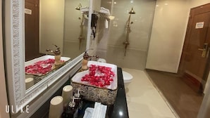 A bathroom with a large mirror, a sink with rose petals, a shower with a golden showerhead, and a toilet. - Hoang Gia I Hotel Yen Xa - by Bay Luxury (Hanoi)
