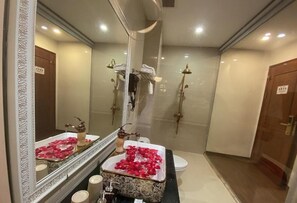 Une salle de bain avec un grand miroir, un lavabo décoré de pétales de rose, une douche dotée d’un pommeau doré et des toilettes.
