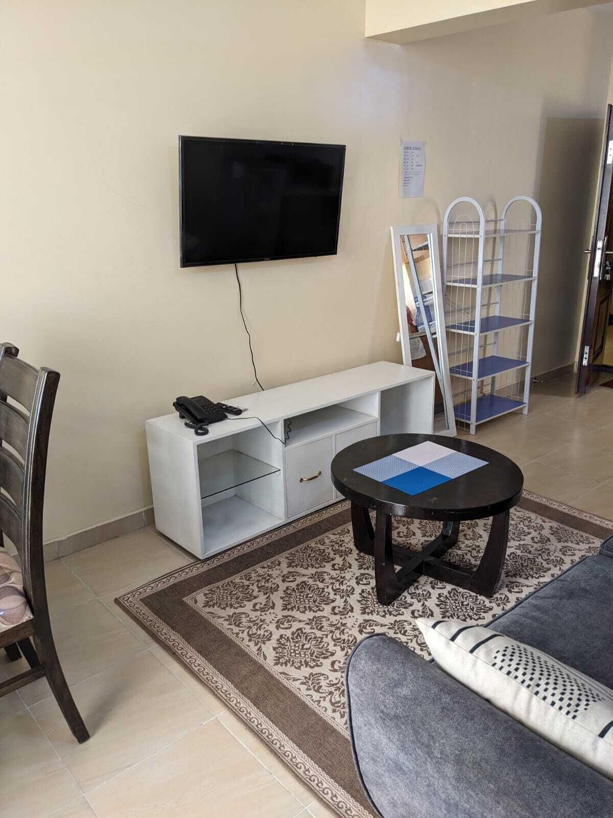Una sala de estar con un televisor de pantalla plana montado en la pared, un mueble blanco para entretenimiento, una mesa de centro redonda con papeles, un sofá gris y una alfombra estampada.