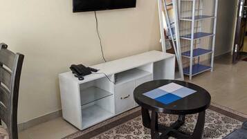 Una sala de estar con un televisor de pantalla plana montado en la pared, un mueble blanco para entretenimiento, una mesa de centro redonda con papeles, un sofá gris y una alfombra estampada.