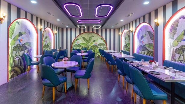 A restaurant with blue chairs and tables, a mural with tropical leaves, and purple neon lights on the ceiling. - Terrazzo Suits (Ahmedabad)