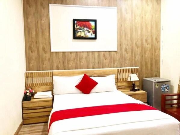 Un lit bien fait, avec un oreiller rouge et une couverture blanche, une tête de lit en bois, une table de chevet avec une lampe, un petit réfrigérateur et un tableau encadré accroché au mur.
