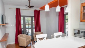 Una sala moderna con piso de madera, una mesa redonda blanca, dos sillones beige, un ventilador de techo y cortinas rojas.