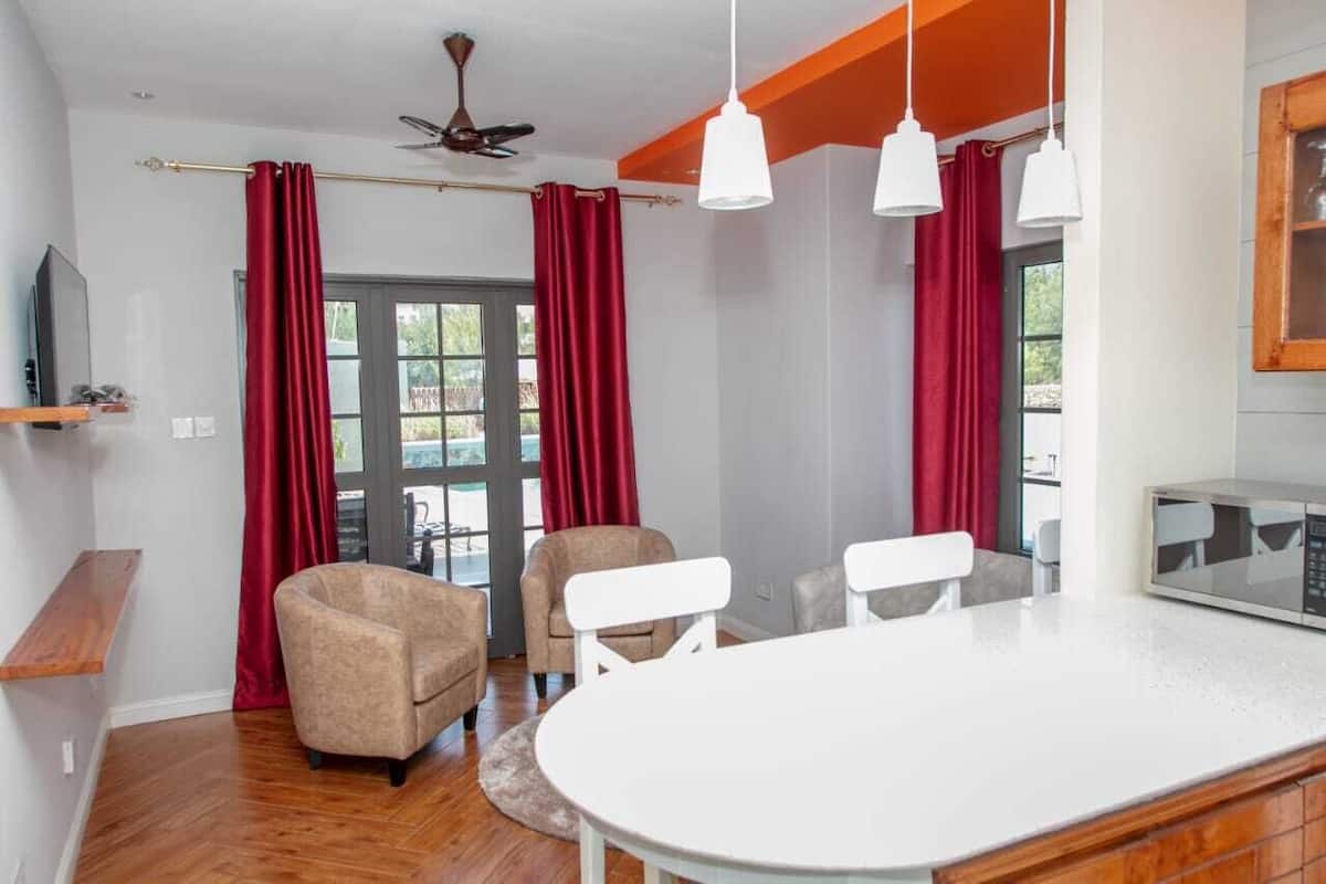 A modern living room with a wooden floor, a round white table, two beige armchairs, a ceiling fan, and red curtains.