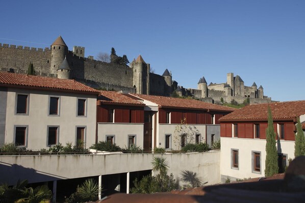 A historic castle with a fortified wall and towers, modern buildings with red shutters in the foreground, and a clear blue sky. - Modern Apartment Near Carcassonne (Carcassonne)