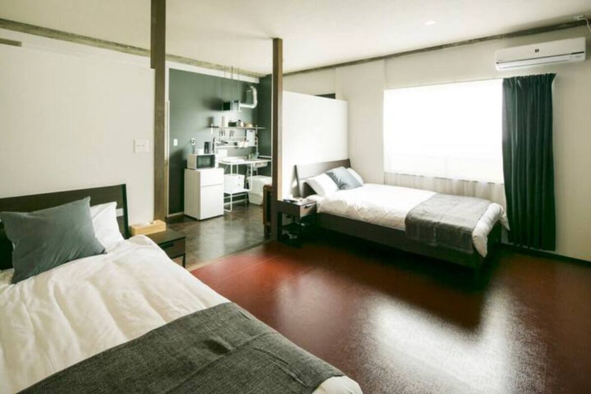 A hotel room with two beds, a kitchenette, a large window with curtains, and an air conditioning unit.
