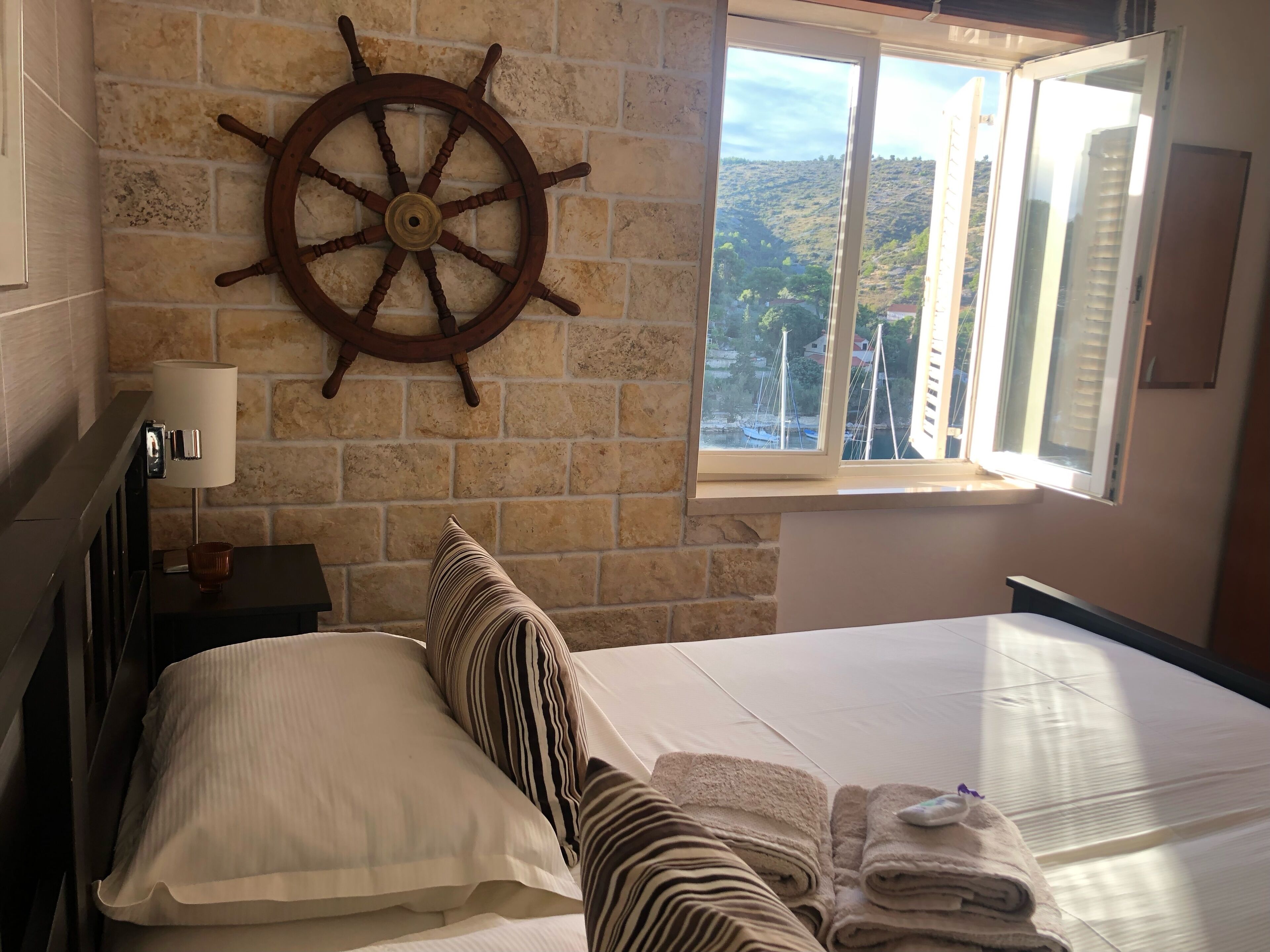 A hotel room with a large bed, a ship's wheel on the wall, a window with a view of the countryside, and a nightstand with a lamp and a cup.