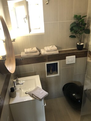 A modern bathroom with a white sink, a mirror, a shelf with folded towels, a potted plant, and a shower area. - Villa More A-11 (Sutivan)