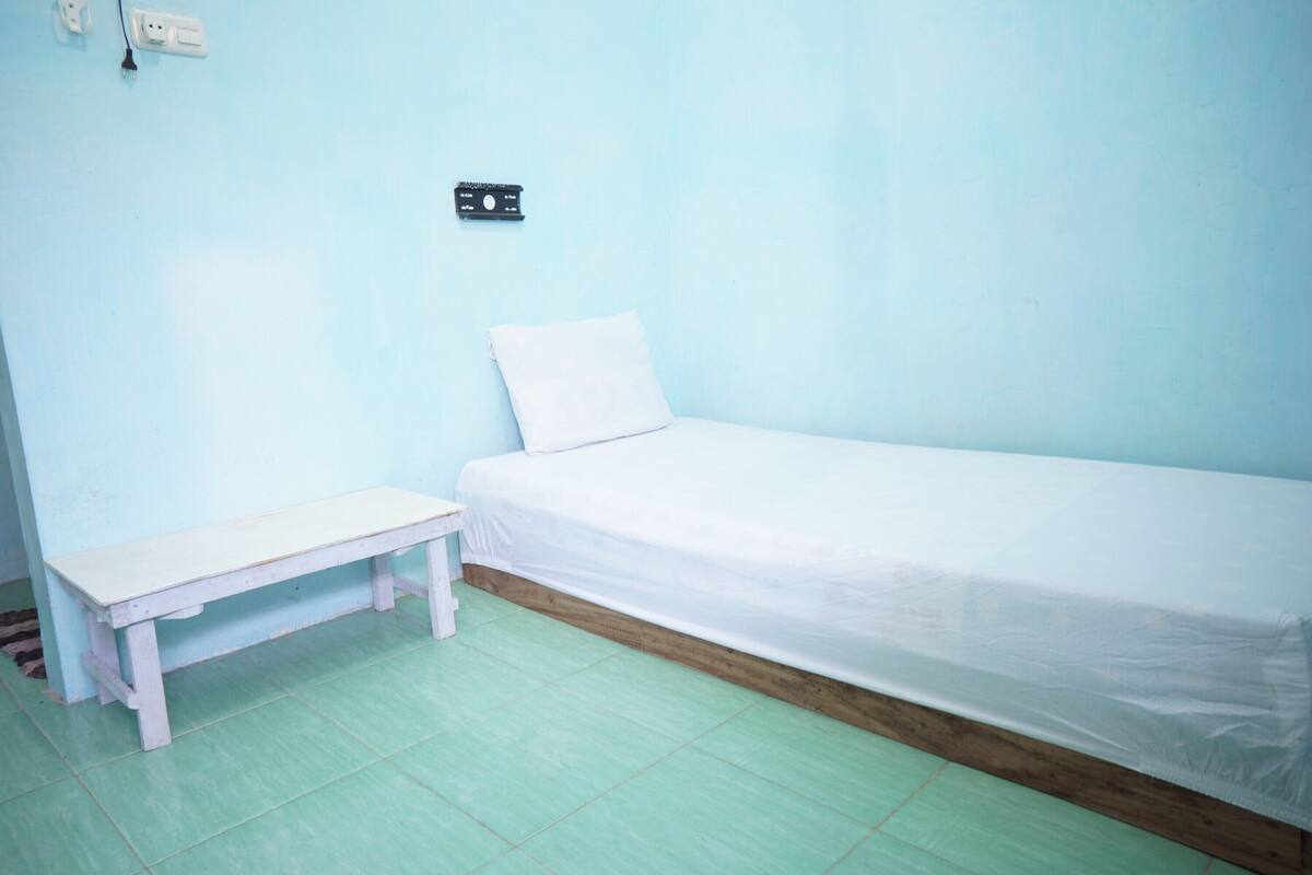 Una habitación pequeña con cama individual, un banco de madera, un almohadón blanco y un enchufe en la pared.
