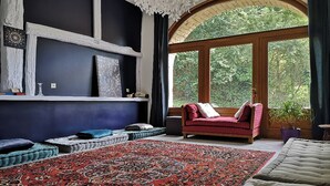 A room with a large red patterned rug, a red sofa with cushions, a wooden chair, and a large arched window with a view of greenery.
