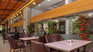 Un restaurante moderno con mesas de madera, asientos tipo banca de cuero, ventanas amplias con plantas y un techo alto.