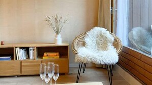 Un salon moderne avec un plancher en bois, un fauteuil blanc moelleux, une table basse en bois sur laquelle reposent trois verres, une bibliothèque et deux lampes fixées au mur.