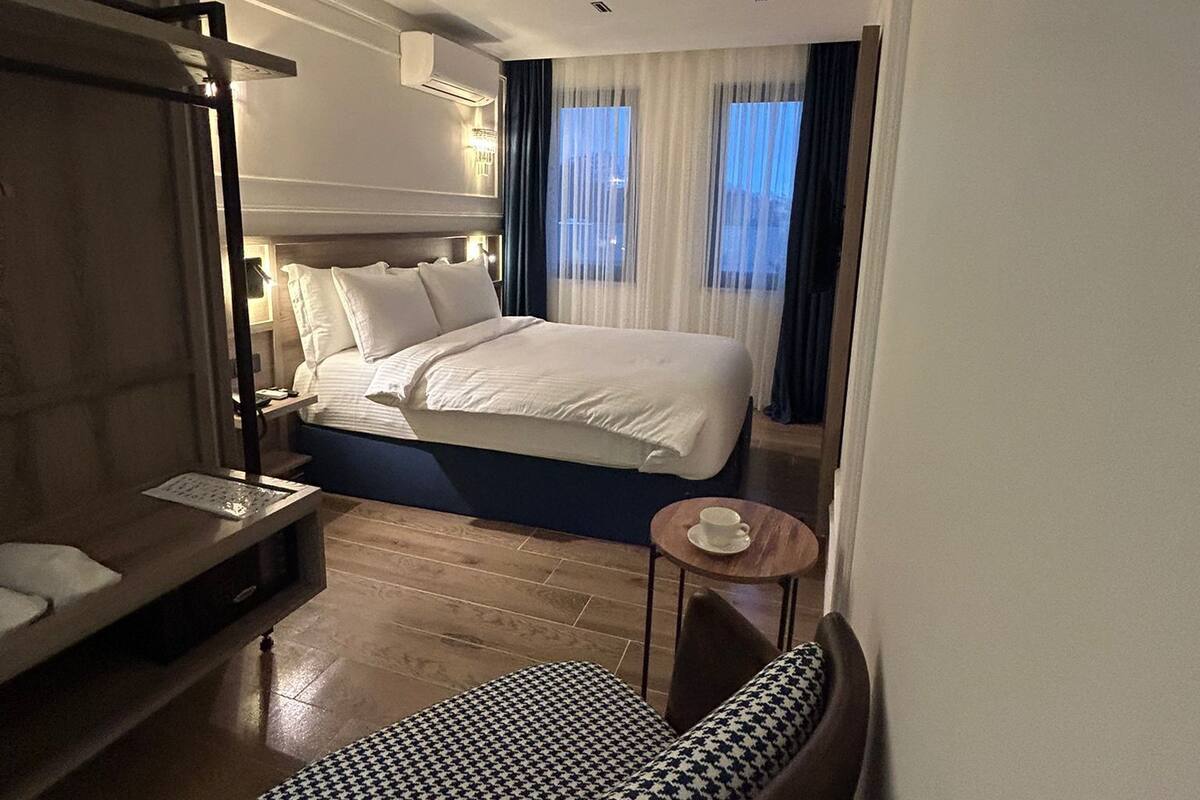 A hotel room with a large bed, a bedside table, a chair, and a small table with a cup.