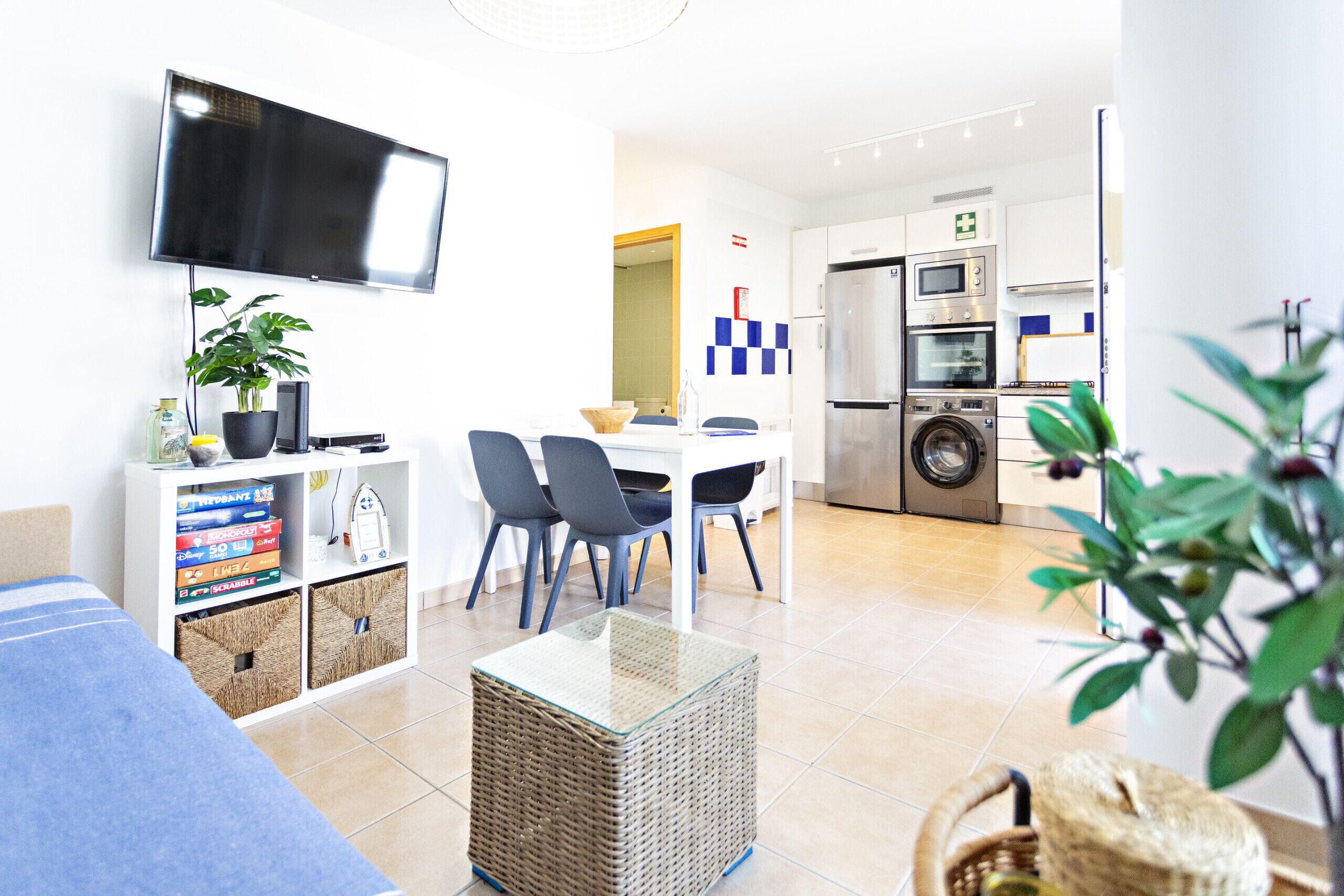 Un salon moderne doté d’un téléviseur à écran plat, d’un coin repas avec des chaises, d’une cuisine équipée et d’un espace buanderie.