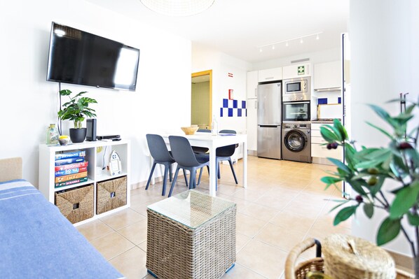 A modern living room with a flat-screen TV, a dining area with chairs, a kitchen with appliances, and a laundry area.