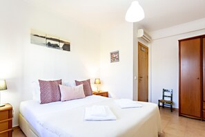 A neatly made bed with white linens and decorative pillows, a bedside table with a lamp, a framed picture on the wall, and a wooden wardrobe.