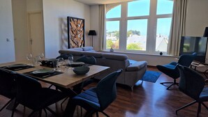 A modern living room with a dining area, large windows, a sofa, and a TV. - Flat in Primel with Sea View & Beaches Nearby (Plougasnou)