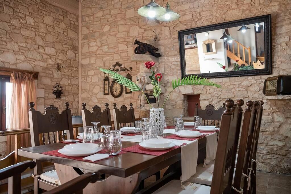 A dining area with a stone wall, wooden chairs, a long table set for four, and a mirror reflecting a staircase.