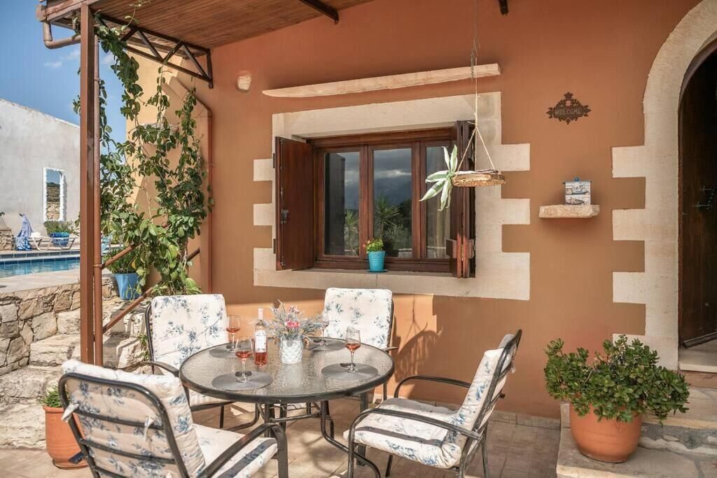 Um pátio externo com uma mesa redonda e quatro cadeiras, rodeado por plantas em vasos e uma pérgola de madeira.