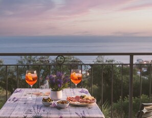 A table with two glasses of orange drink, a plate of food, and a potted plant on a balcony overlooking a body of water. - Casa Conca Azzurra (Costarainera)