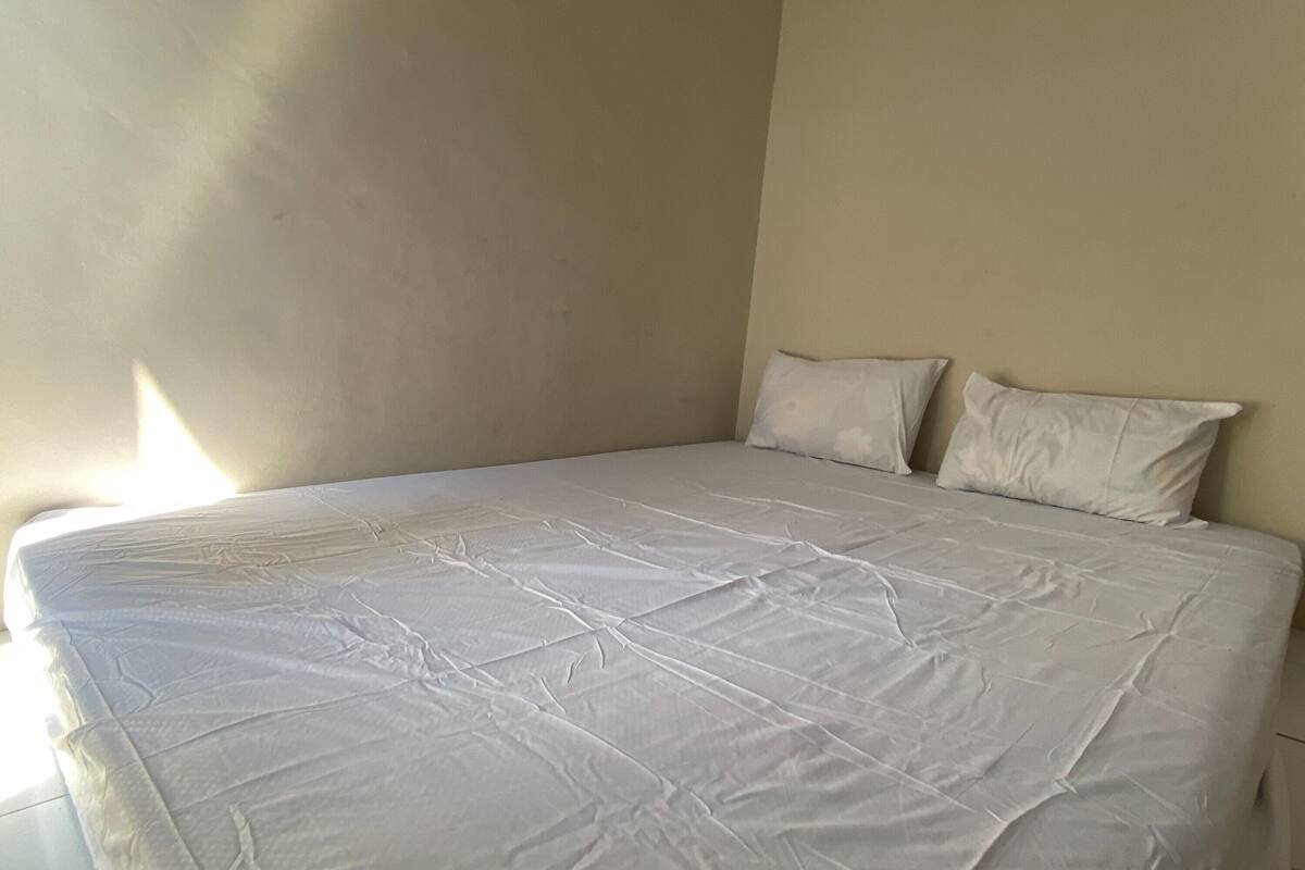 Una cama con sábana blanca y dos almohadas en una habitación con paredes lisas y un ventanal.