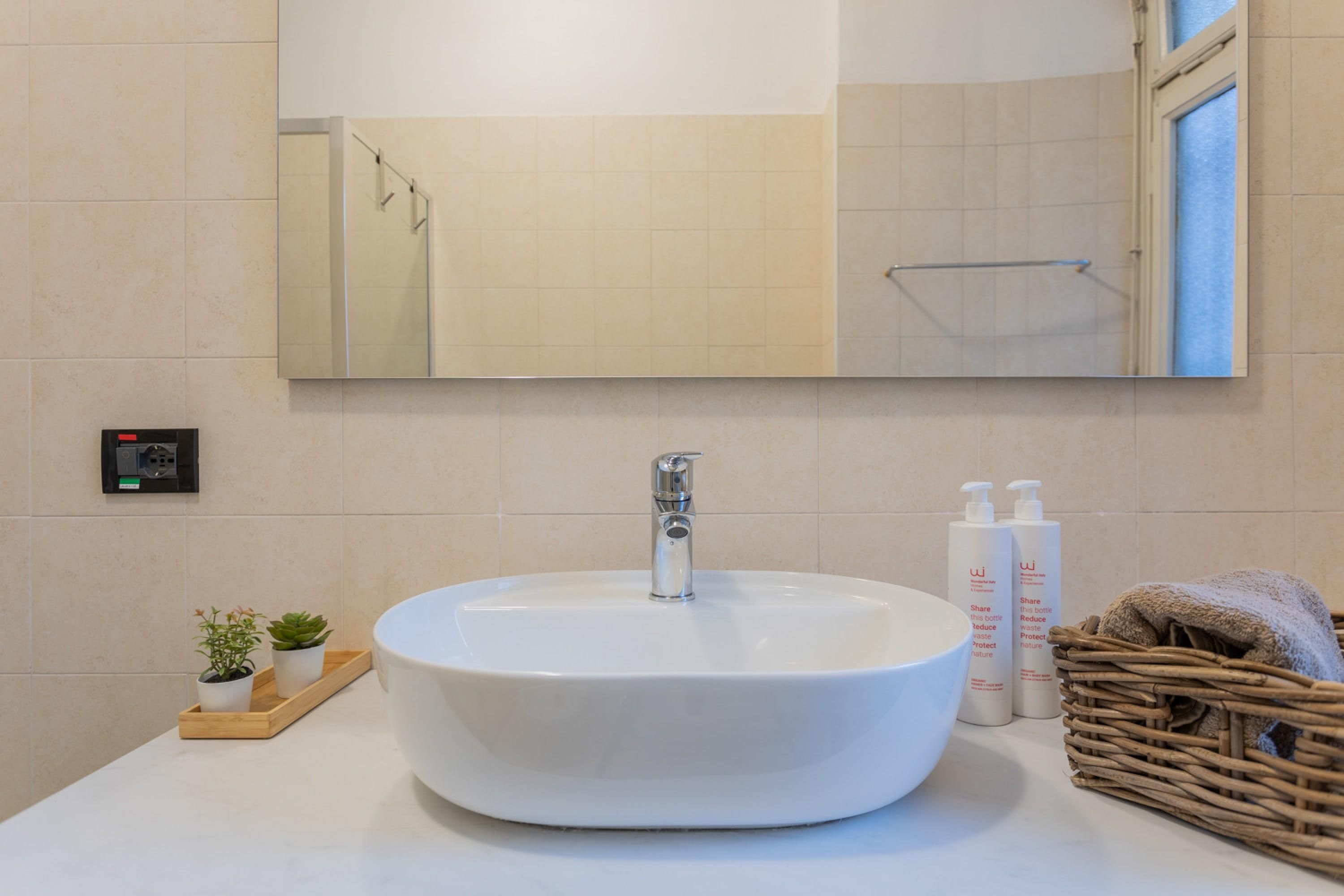 A bathroom with a round white sink, a mirror above it, and a window to the right. There are two potted plants on the counter and a wicker basket with towels.