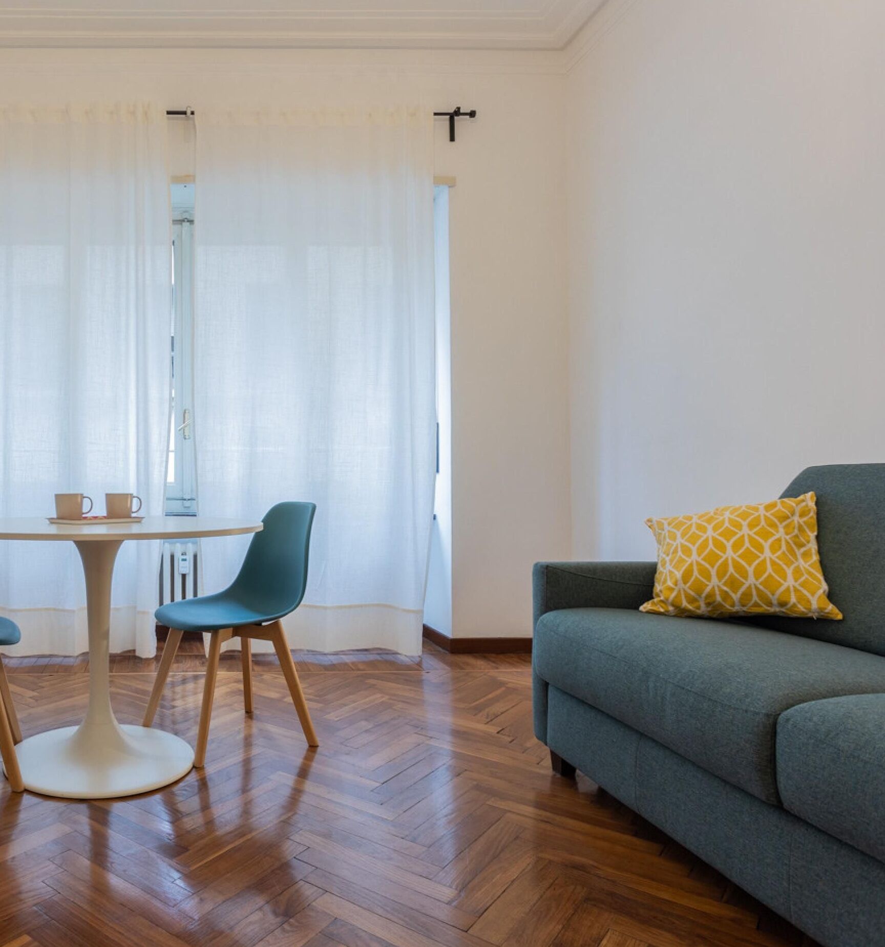 Un salon avec un canapé vert, un coussin jaune à motifs, une table avec des chaises et une fenêtre ornée de rideaux légers.