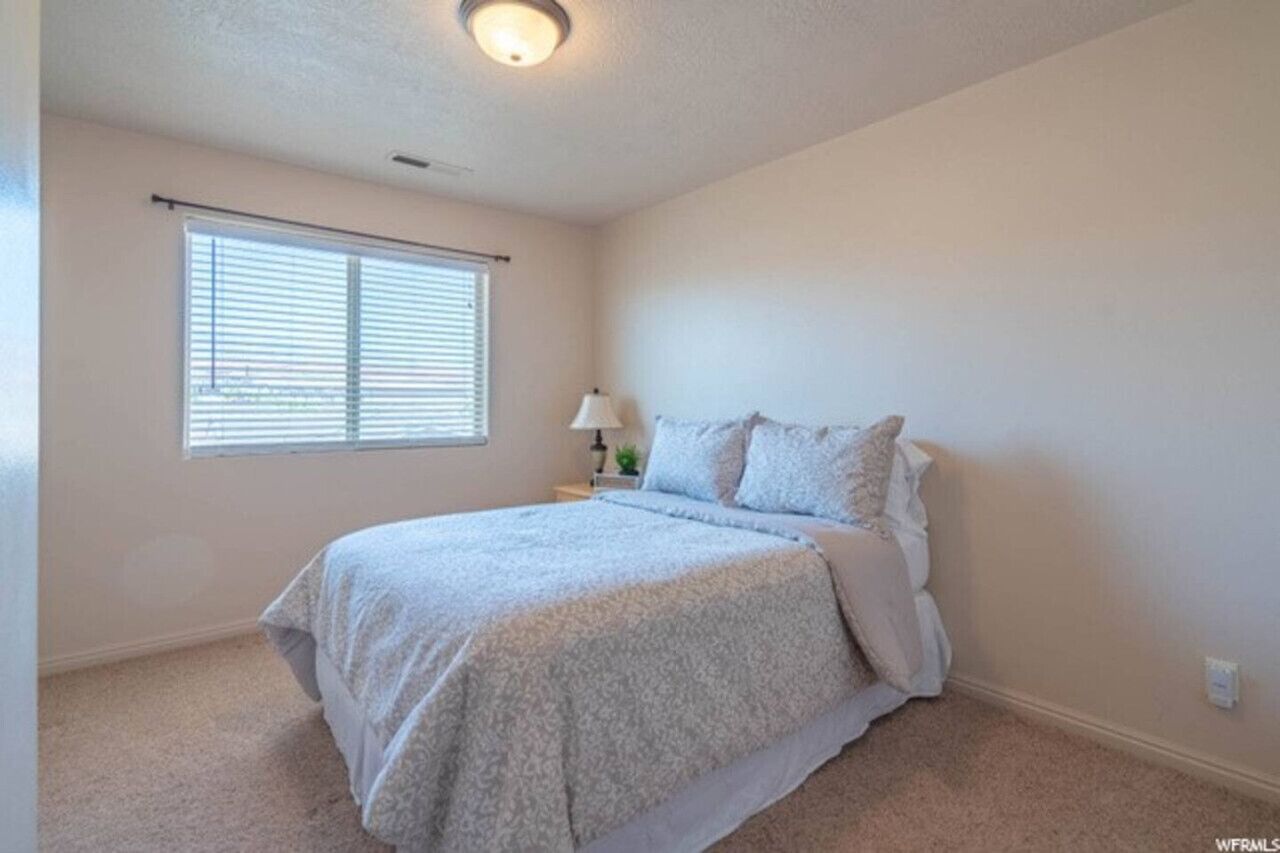 Une chambre à coucher avec un lit, une fenêtre avec des stores, une table de chevet avec une lampe et un luminaire au plafond.