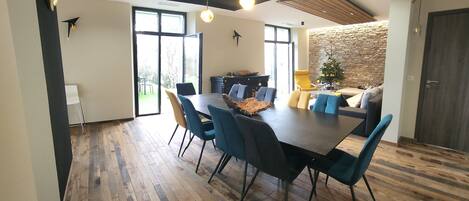 A modern dining area with a wooden floor, a large table, and blue chairs. There is a stone wall and a door in the background.