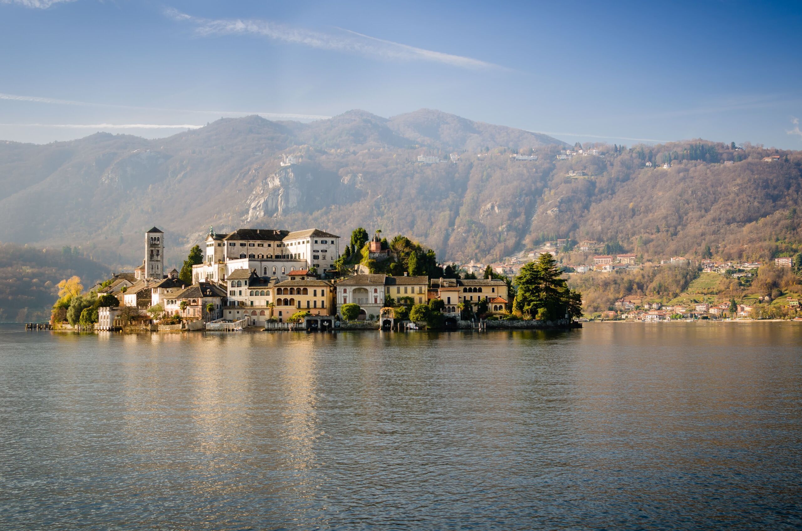 Un edificio junto al lago con varios pisos, una torre destacada y montañas al fondo.
