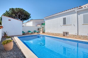 A swimming pool with a clear blue water, surrounded by a white building with a stone base, and a tree in the background.
