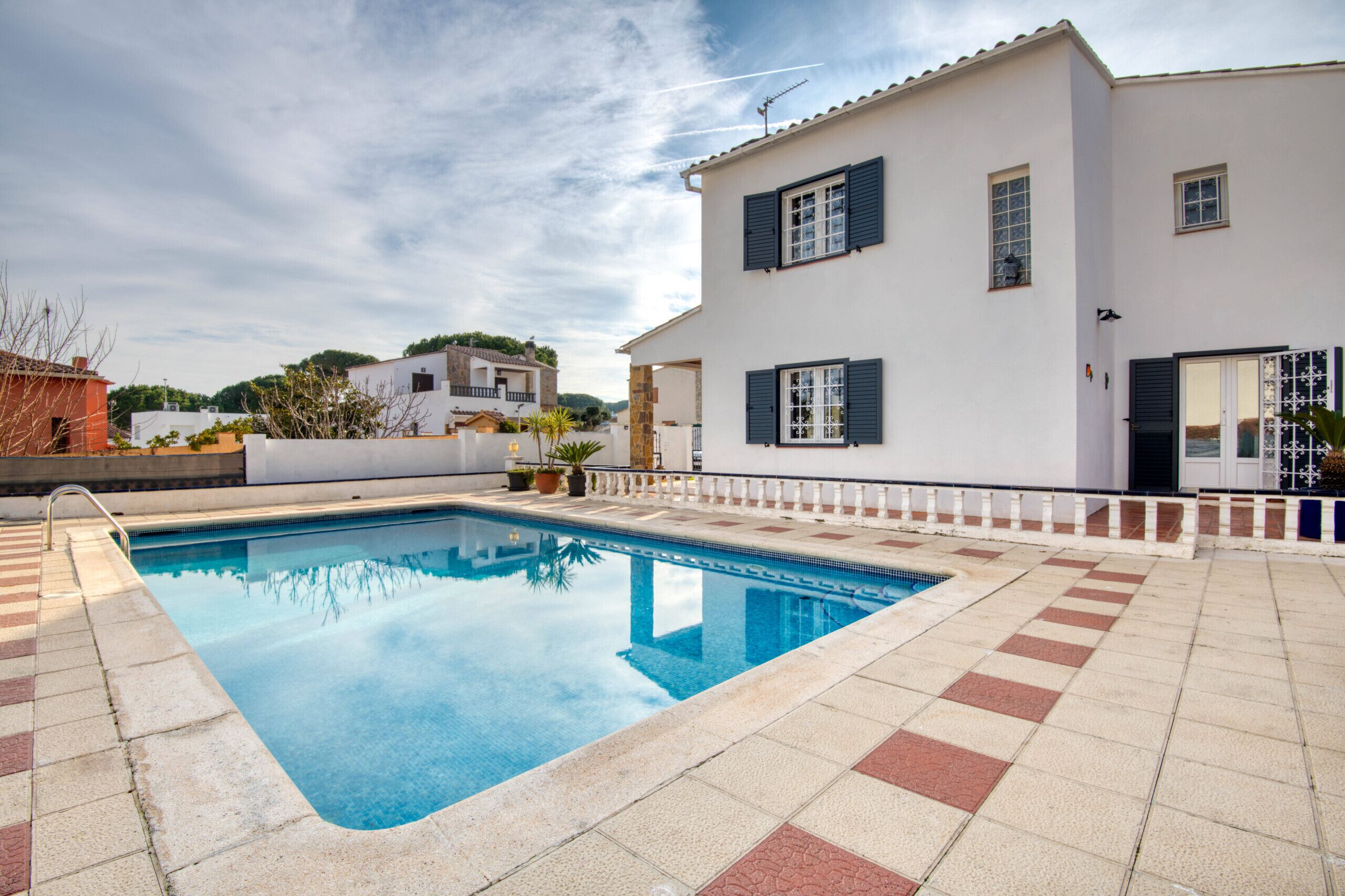 A swimming pool with a tiled deck, a white building with dark shutters, and a clear sky.
