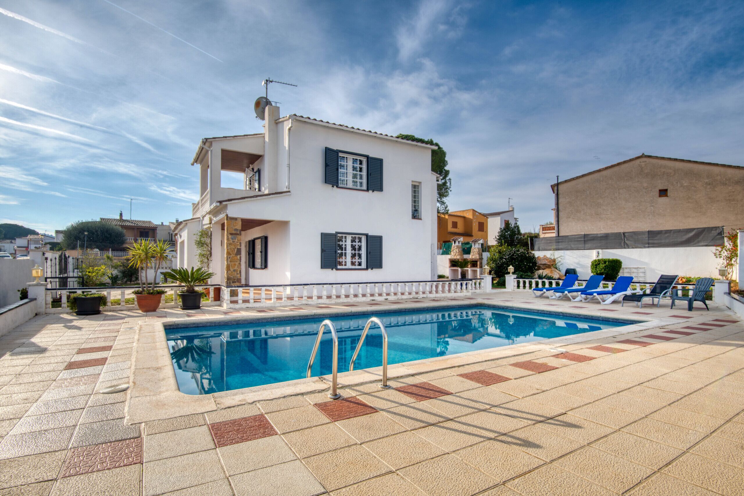 Una piscina amb una terrassa enrajolada, un edifici blanc amb persianes negres i un cel blau clar.